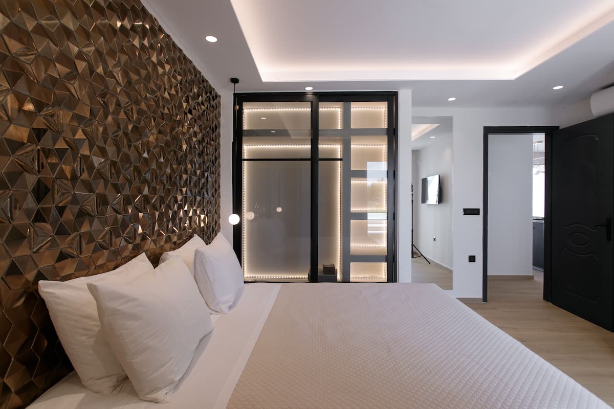 A modern bedroom with a geometric patterned headboard, a large bed with white linens, and a sliding glass door leading to a bathroom.
