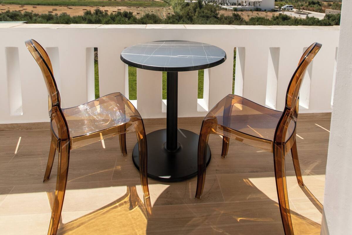 A balcony with a round table and two clear chairs, overlooking a landscape with distant buildings and mountains.