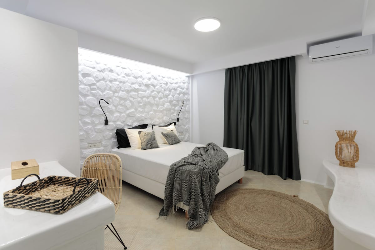 A modern bedroom with a white bed, a textured wall, a large window, a woven chair, and a woven rug.