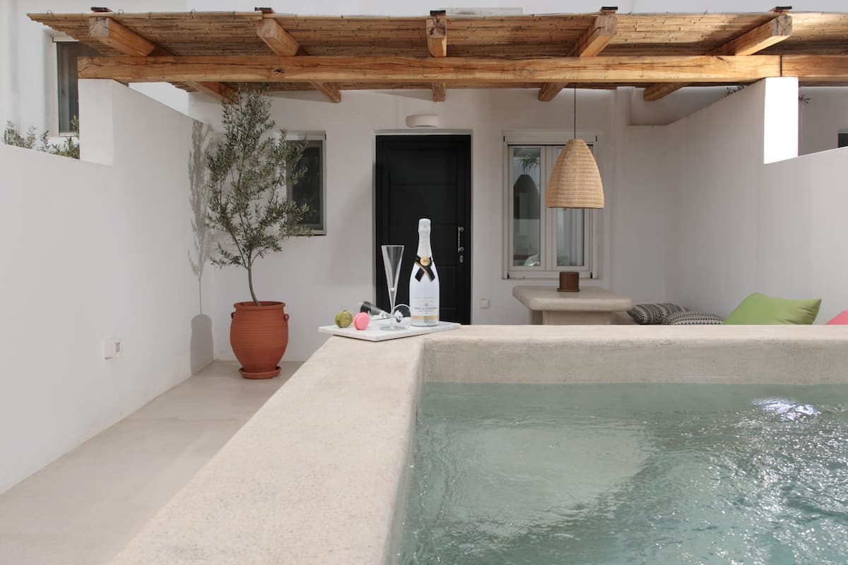 A pool area with a wooden pergola, a potted plant, and a small table with a bottle and glasses.