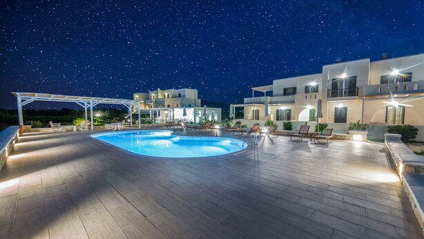 Una piscina con sillones de descanso y una zona cubierta, rodeada de edificios modernos con iluminación exterior.