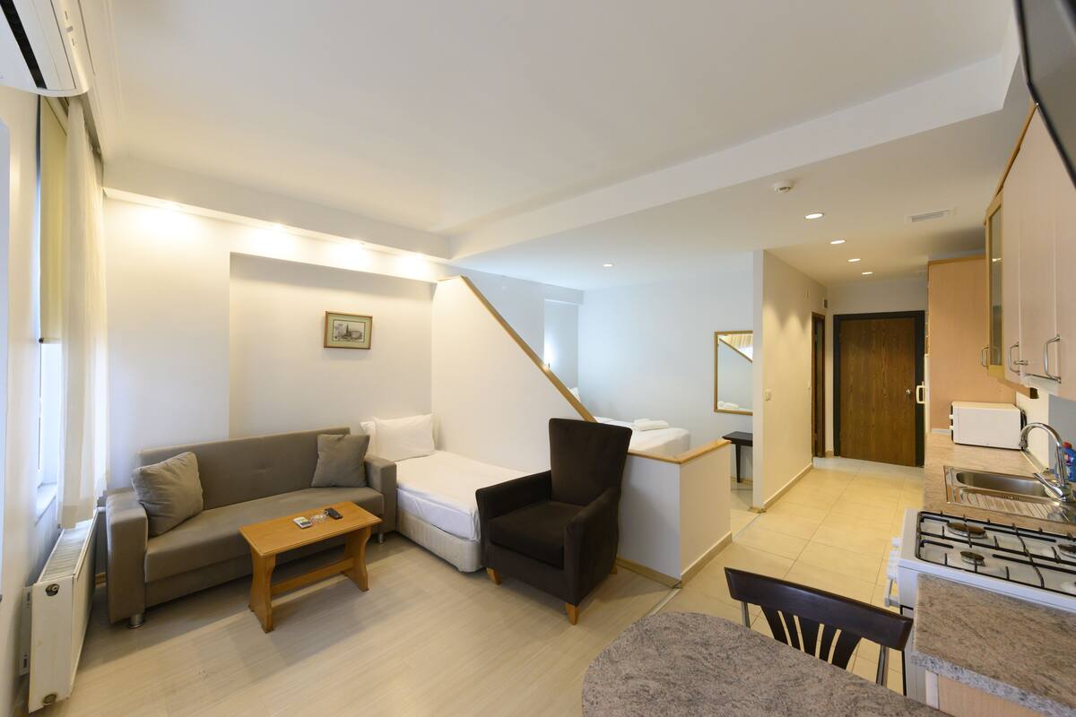A compact living space with a sofa, armchair, and a small dining table. A kitchenette with a stove and sink is visible.