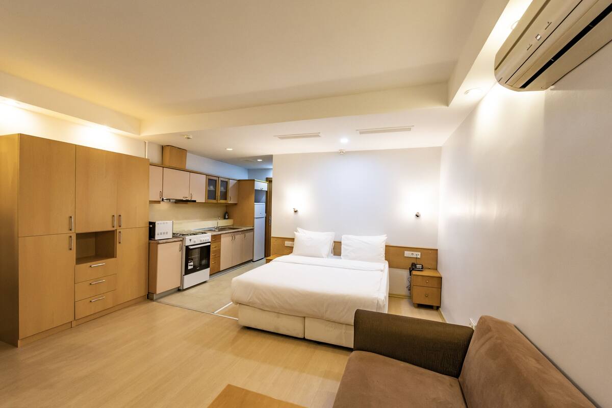 A modern hotel room with a large bed, a sofa, a kitchenette, and a separate bathroom.