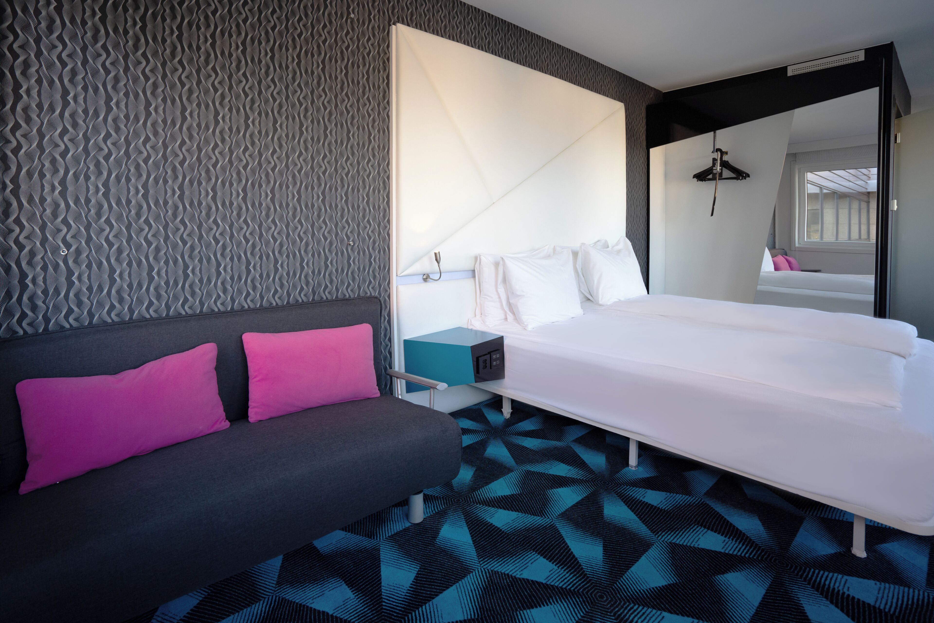 Una habitación de hotel moderna con una cama grande, un sofá con cojines rosados, un suelo con estampado geométrico y una pared con textura original.