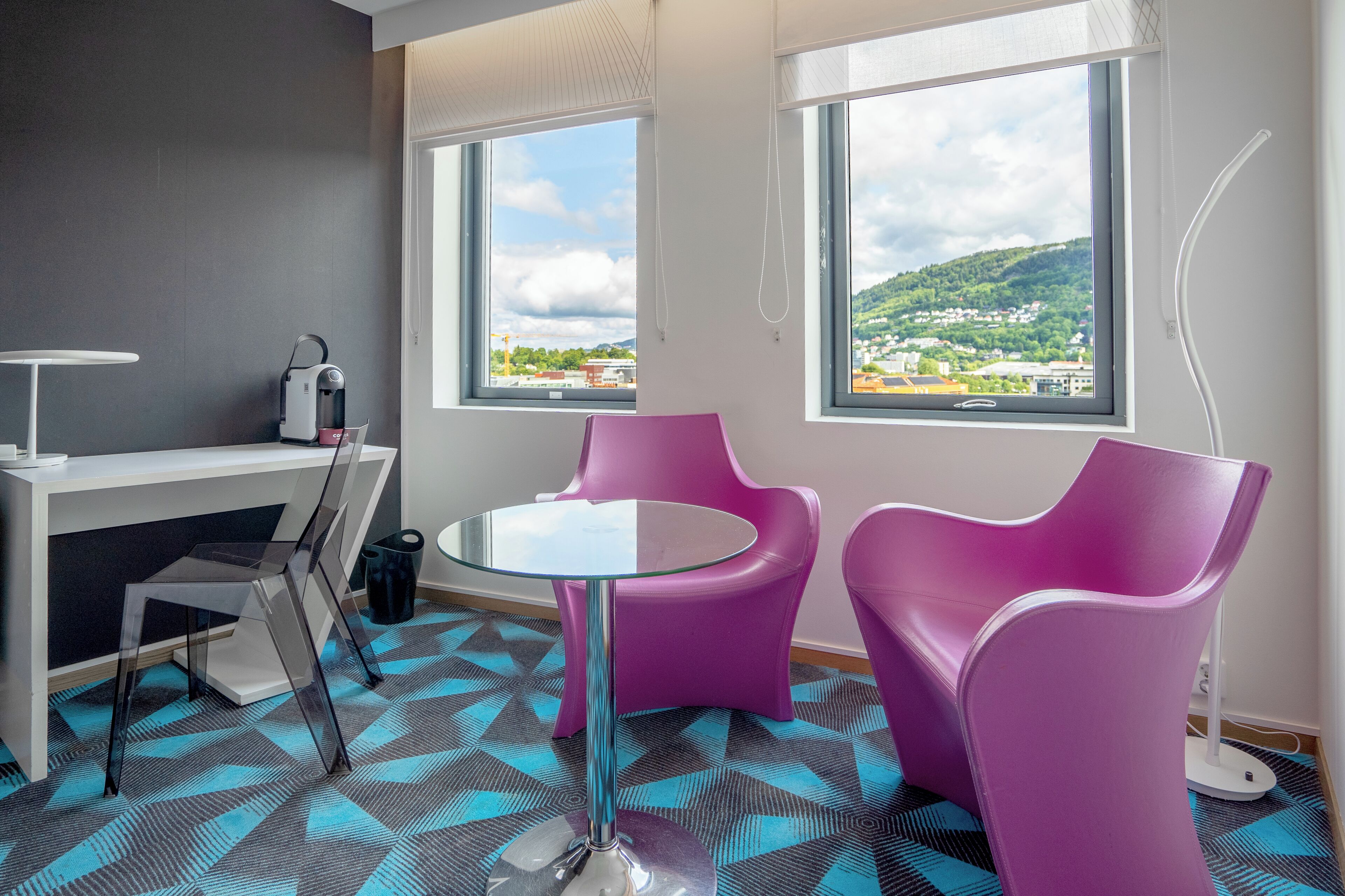 A modern room with a desk, two purple chairs, a glass table, and large windows offering a view of a green landscape.