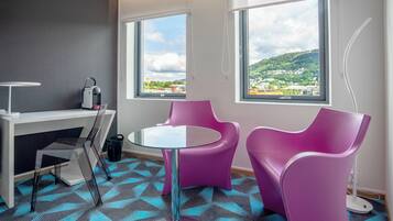 A modern room with a desk, two purple chairs, a glass table, and large windows offering a view of a green landscape.
