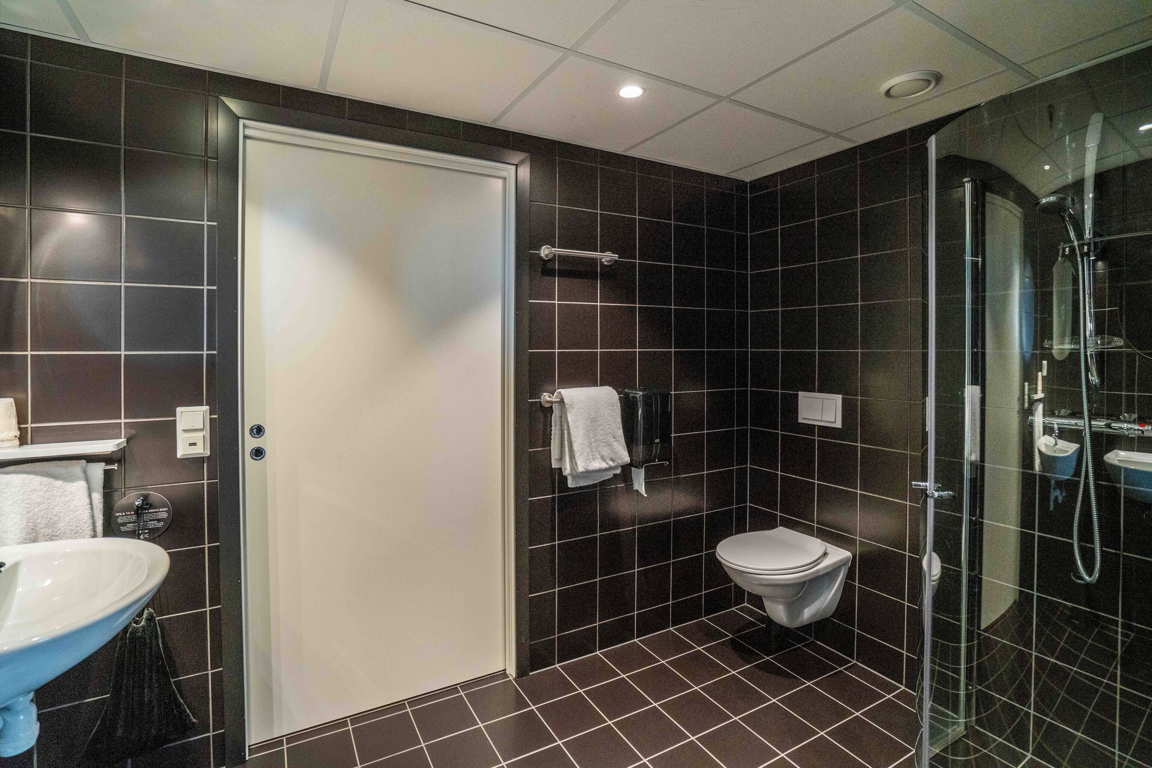 Un baño moderno con un inodoro blanco, ducha con mampara de vidrio y lavamanos con toallero.