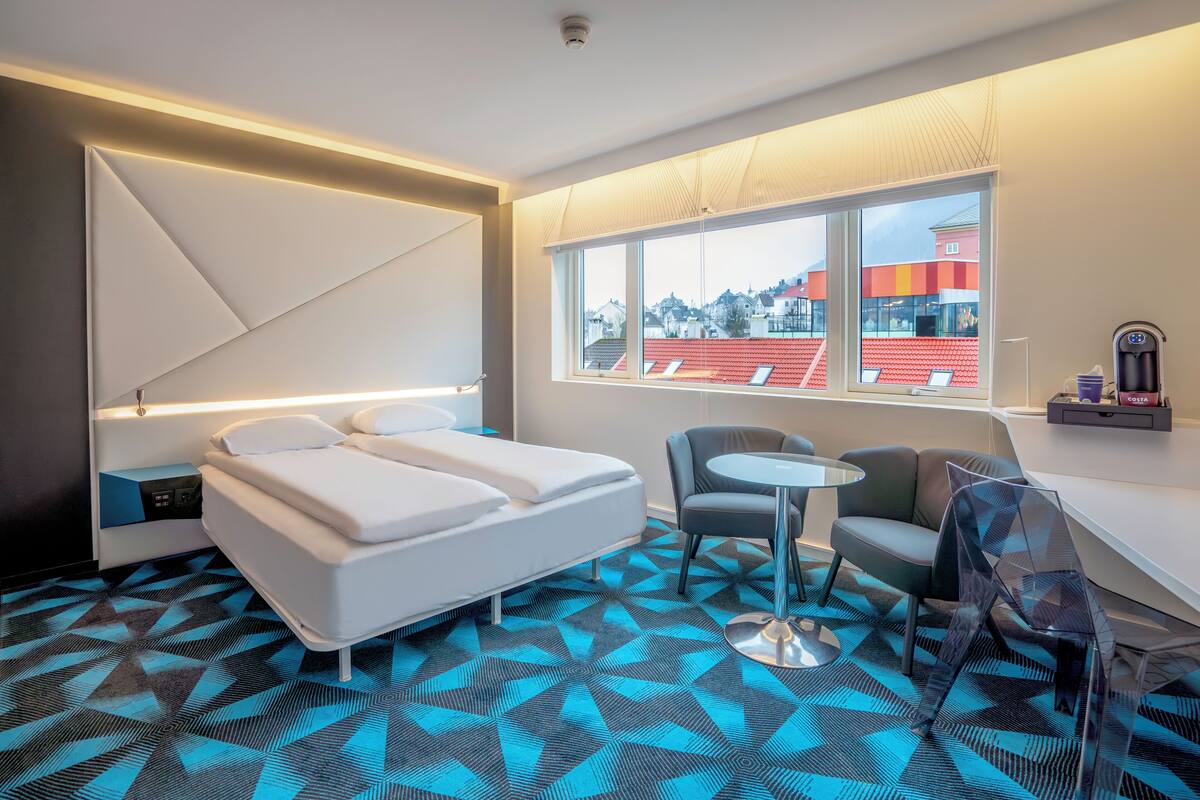 A modern hotel room with a bed, a small table, a chair, and a large window offering a view of buildings outside.