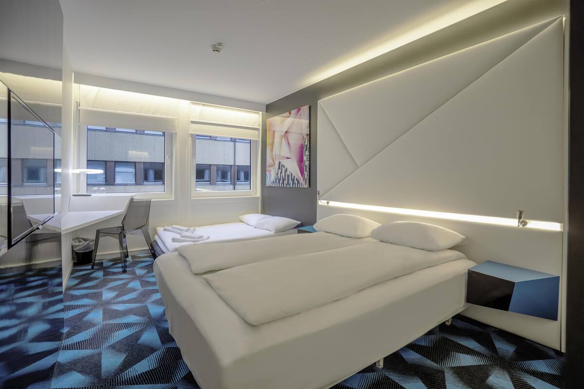 A modern hotel room with a bed, desk, chair, and large windows.