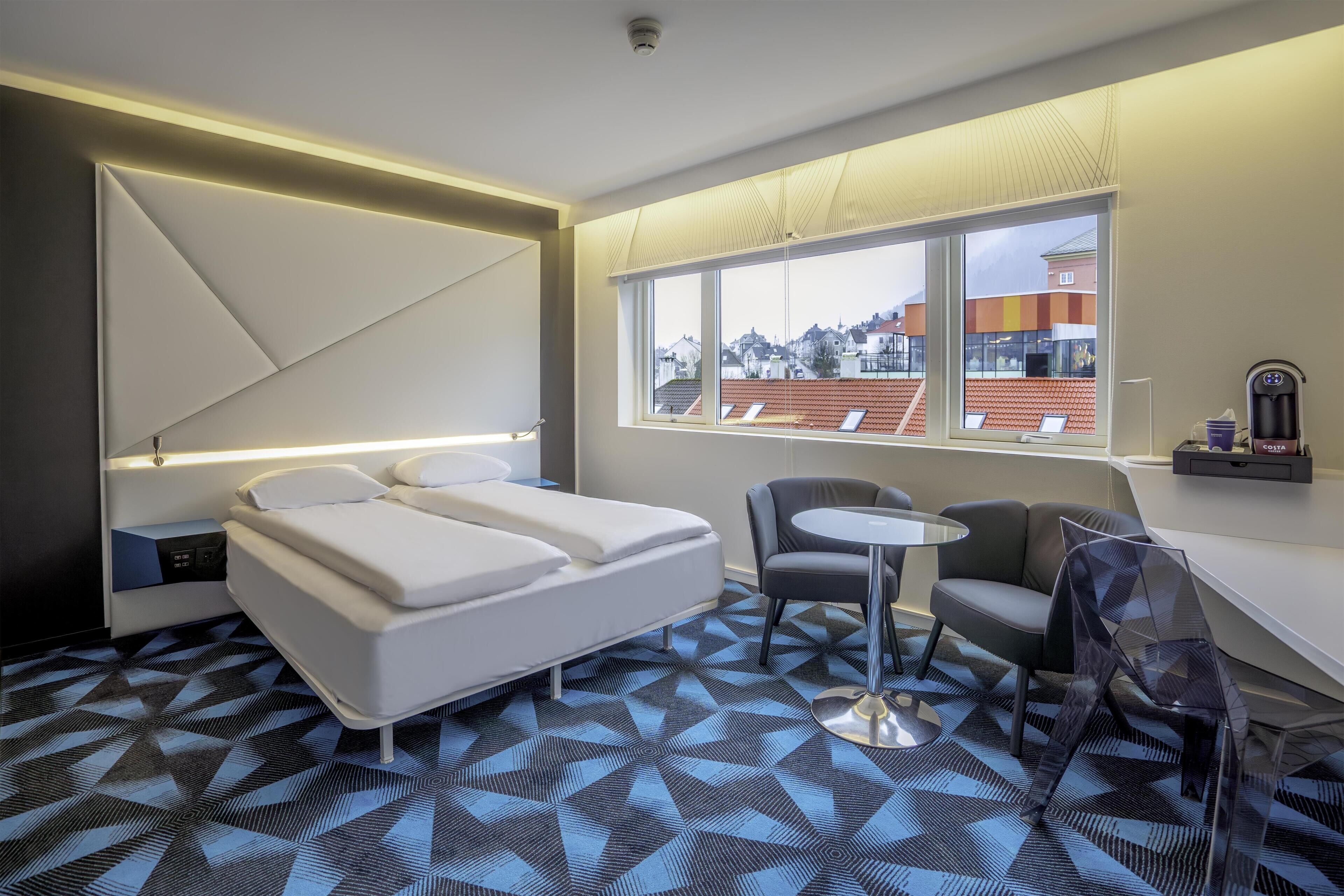 Una habitación de hotel moderna con cama, una mesita, una silla y una ventana con vistas a edificios.