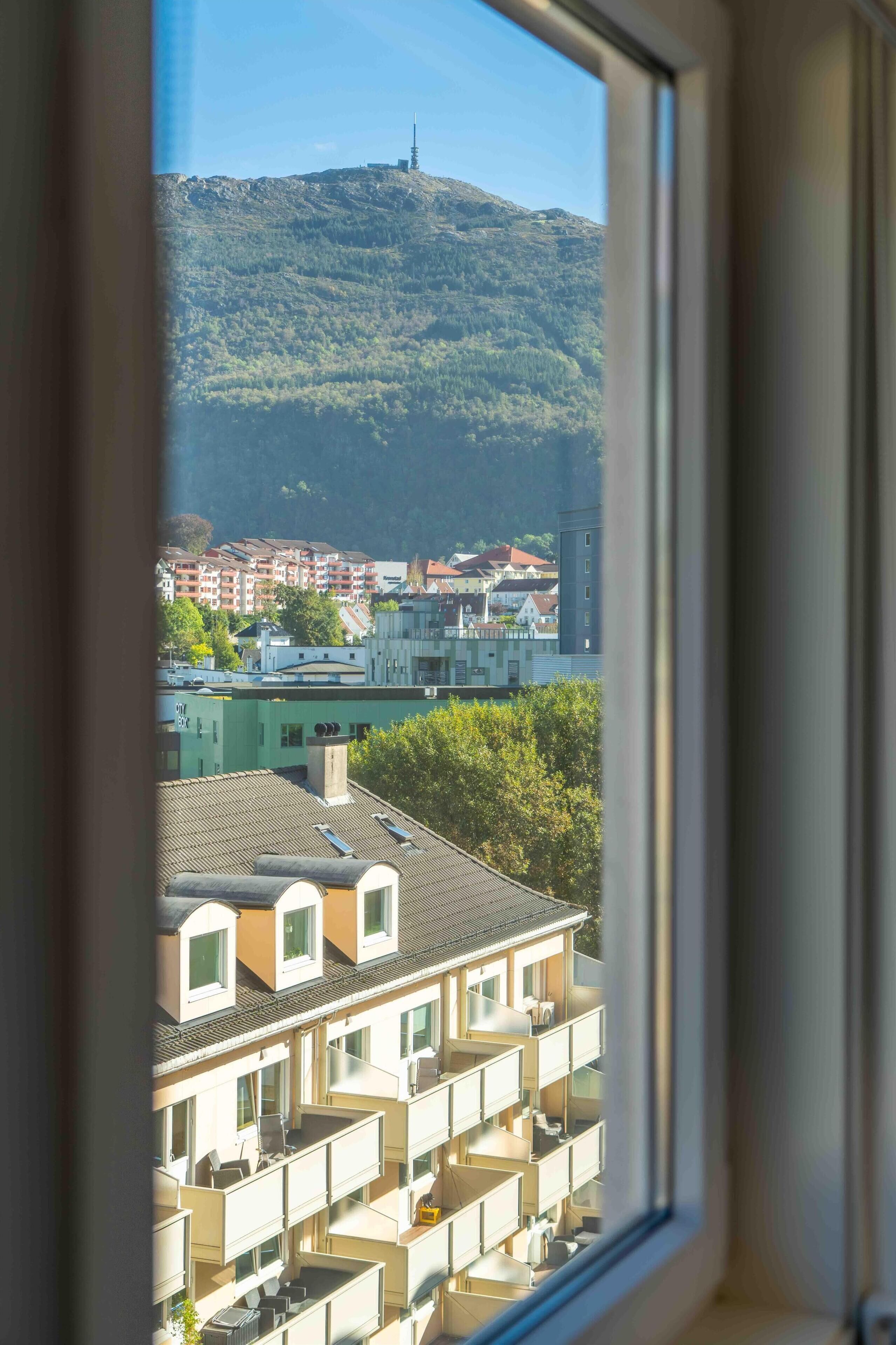 A view through a window of a building with multiple balconies, a mountain in the background, and a distant tower.