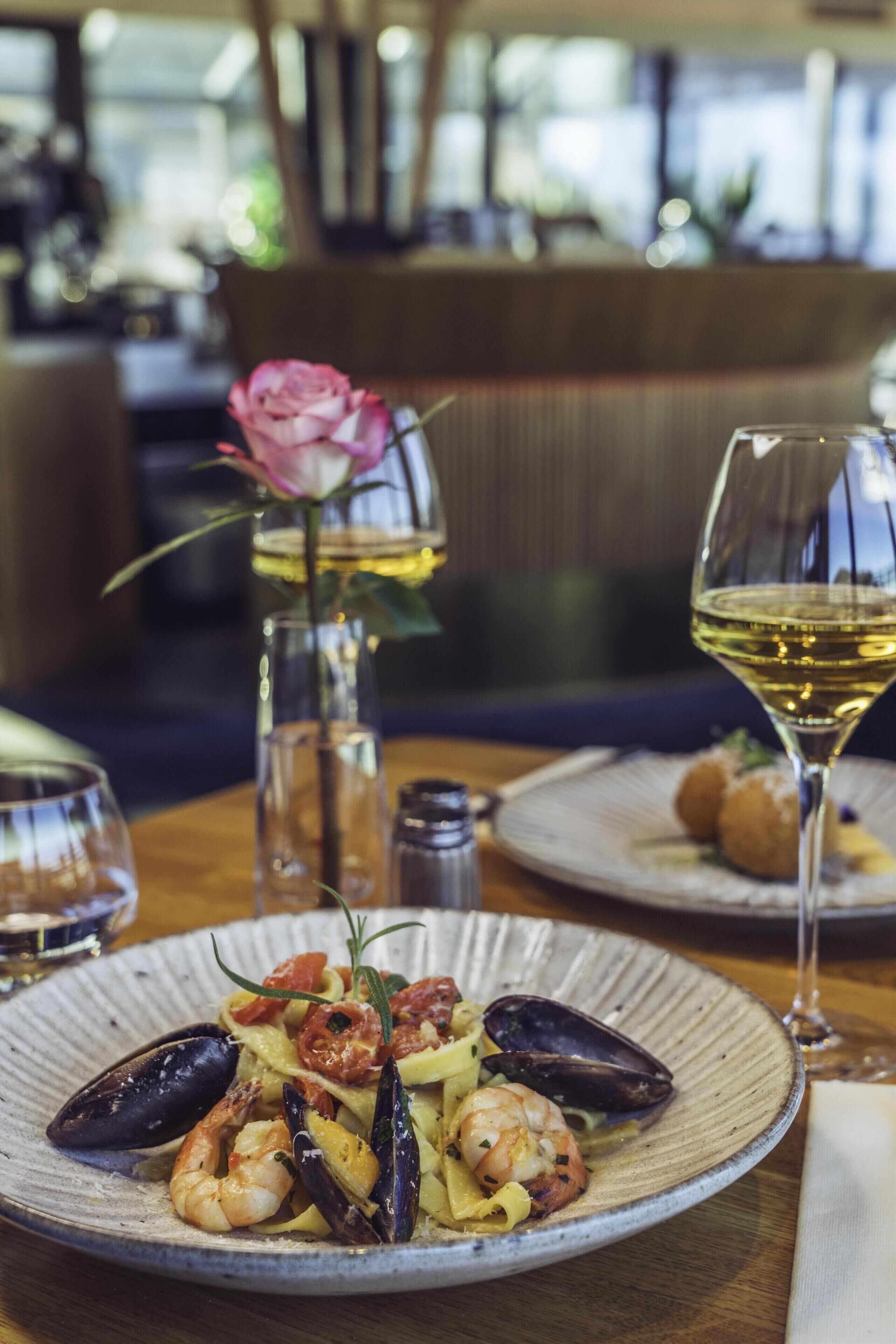 A dining table with plates of seafood pasta, a glass of white wine, and a vase with a pink flower.