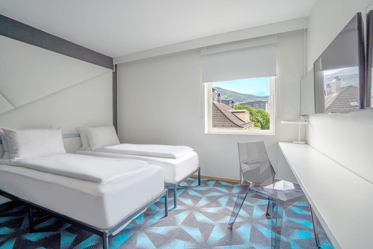 A modern hotel room with two beds, a desk, and a window with a view of buildings and hills.