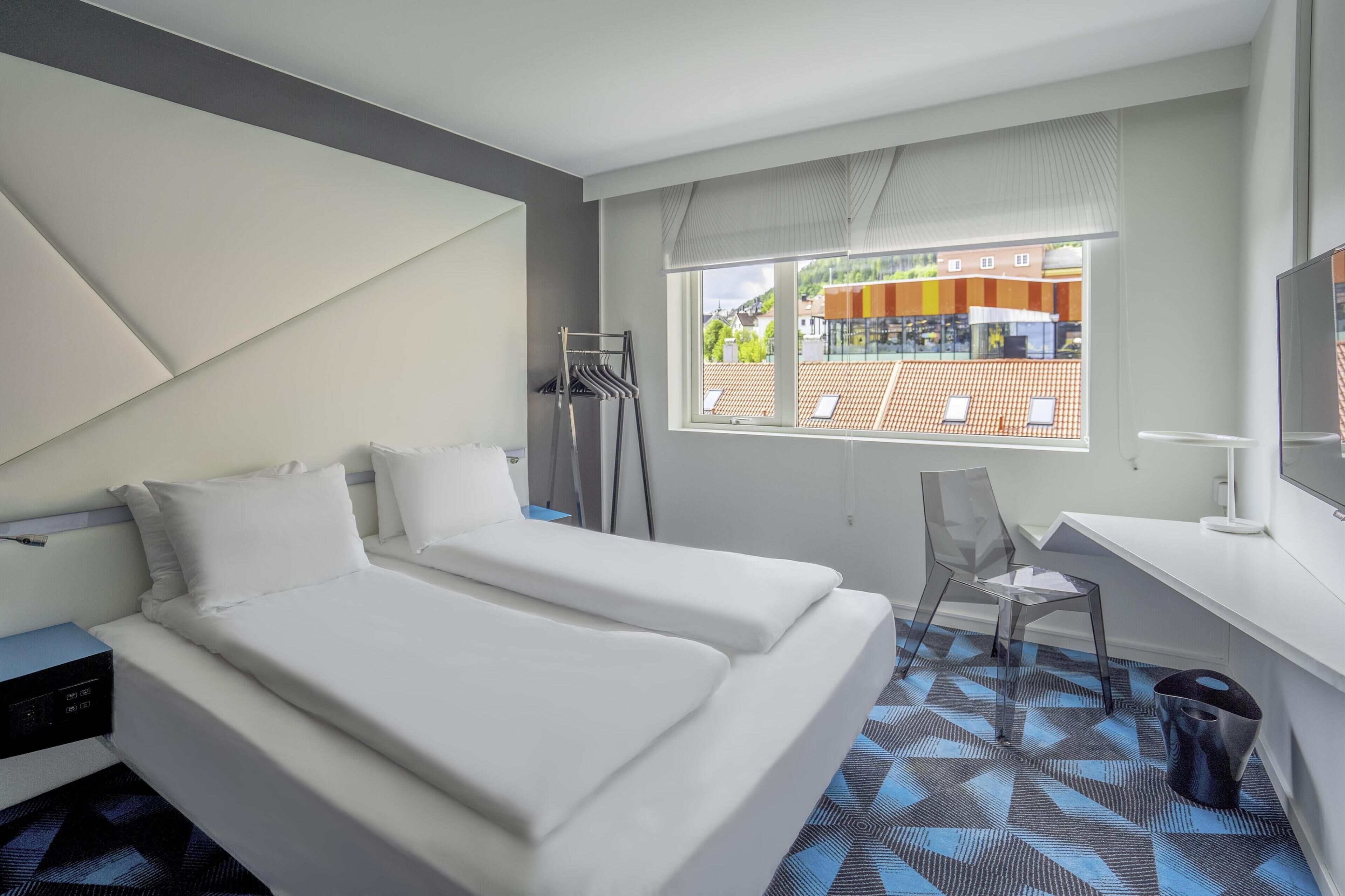 Et moderne hotelværelse med to senge, et skrivebord, en stol, et fjernsyn og et vindue med udsigt til bygninger.