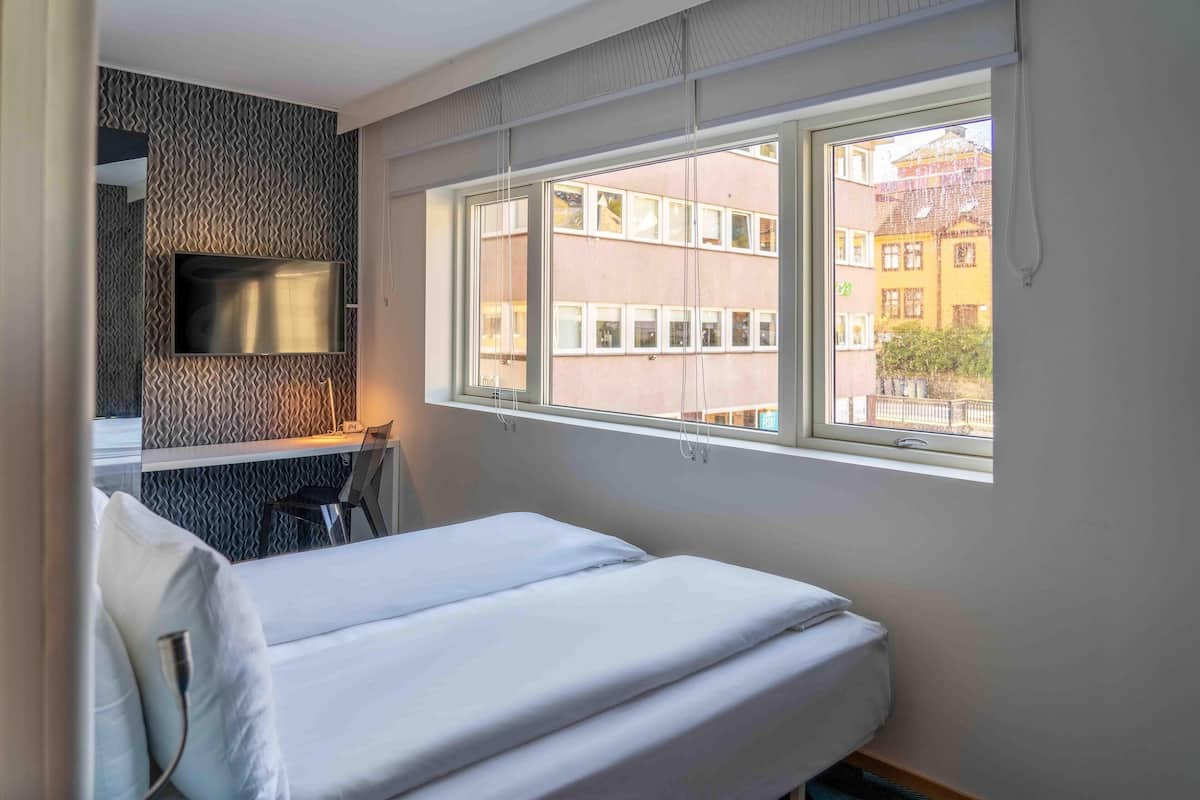 A hotel room with a bed, a TV mounted on the wall, and a window with a view of buildings.