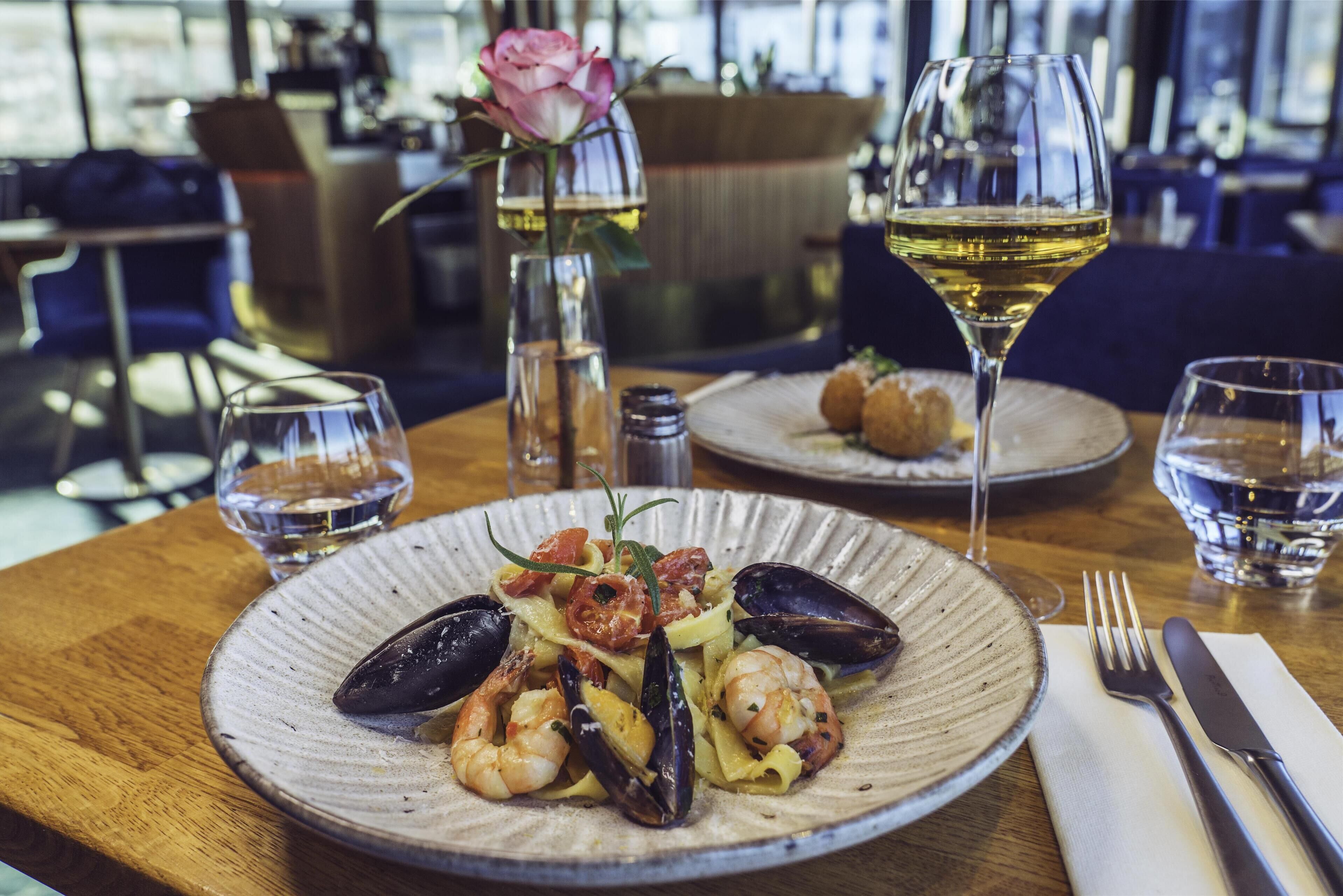 A seafood dish on a plate with a glass of white wine and a rose in a vase.