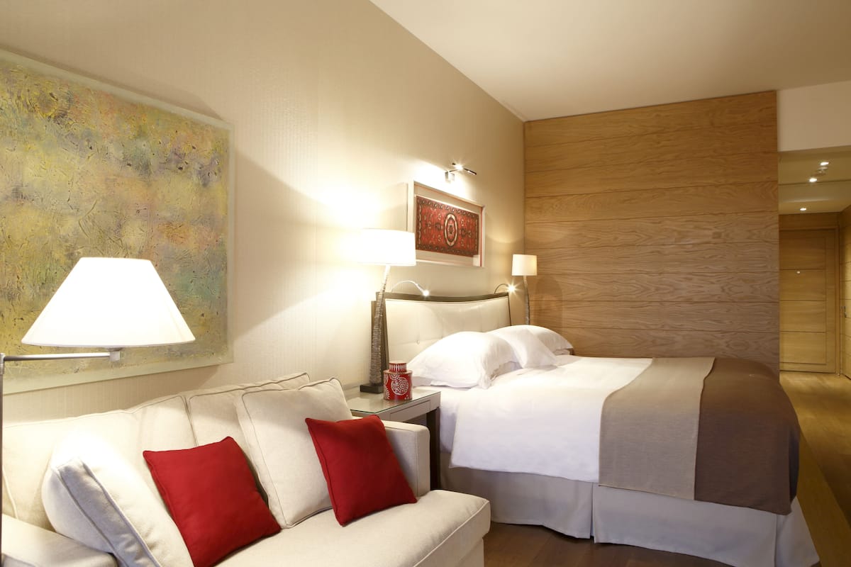 A hotel room with a bed, a sofa, a nightstand, a lamp, and a painting on the wall.