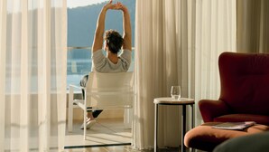 A room with a large window, a red chair, a small table, and a person stretching. - Chenot Palace Gabala (Gabala)