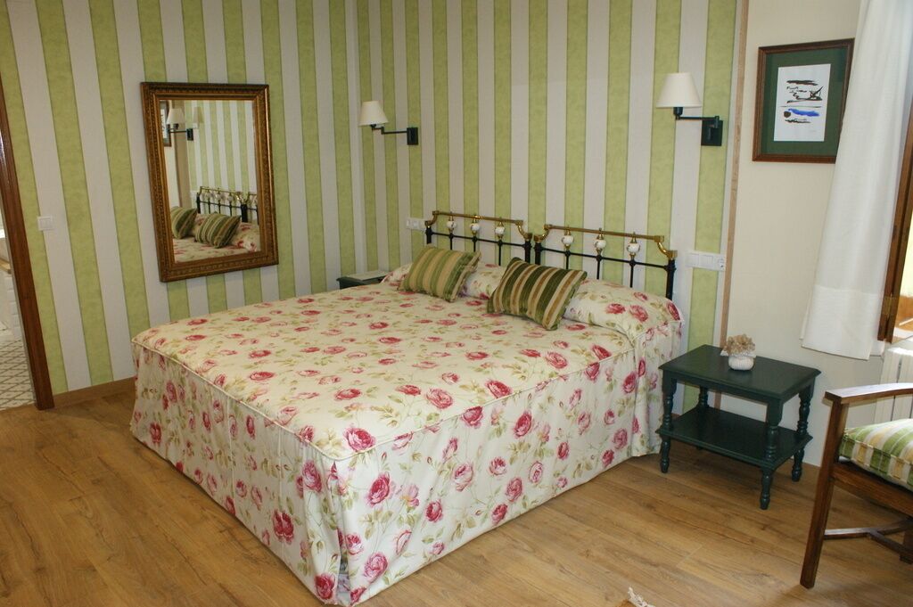 A bedroom with a striped green and white wall, a bed with floral bedding, a nightstand, a chair, a mirror, and a framed picture on the wall.
