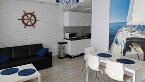 A modern living area with a white brick wall, a black sofa, a white dining table with chairs, a blue pendant light, and a kitchen area with white cabinets and a microwave. - Family Homes - Apartamenty Rzemieślnicza (Władysławowo)