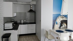 A compact kitchen with white cabinets, a black countertop, and a tiled backsplash. There's a small sink and a kettle on the counter. A dining area with a glass table and white chairs is visible, featuring a large window with a view of the sea. - Family Homes - Apartamenty Rzemieślnicza (Władysławowo)