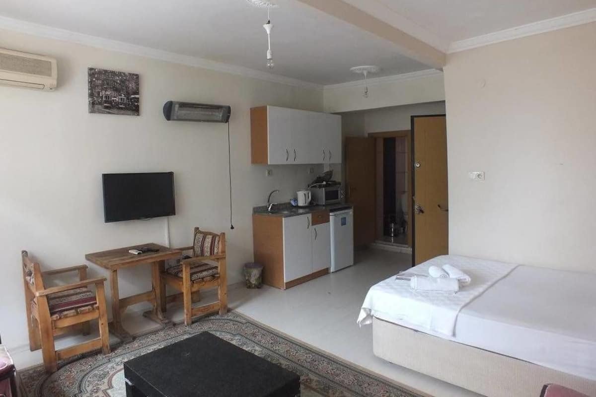 A compact room with a bed, a small kitchenette, a dining area with a table and chairs, a TV, and a ceiling fan.