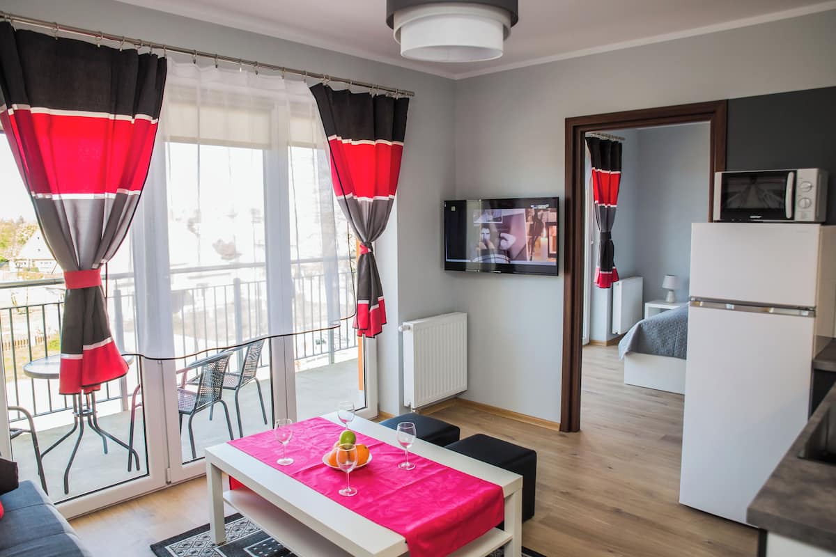 Ein modernes Hotelzimmer mit Balkon, einer Essecke mit Tisch für zwei Personen, einer Küchenzeile und einem an der Wand befestigten Fernseher.