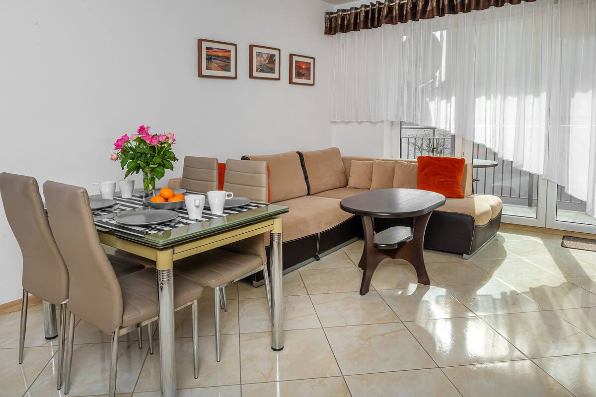Ein modernes Wohnzimmer mit Essbereich, ausgestattet mit einer Couch, Sesseln, einem Couchtisch und einem Esstisch, gedeckt mit Tellern, Tassen und einer Blumenvase.
