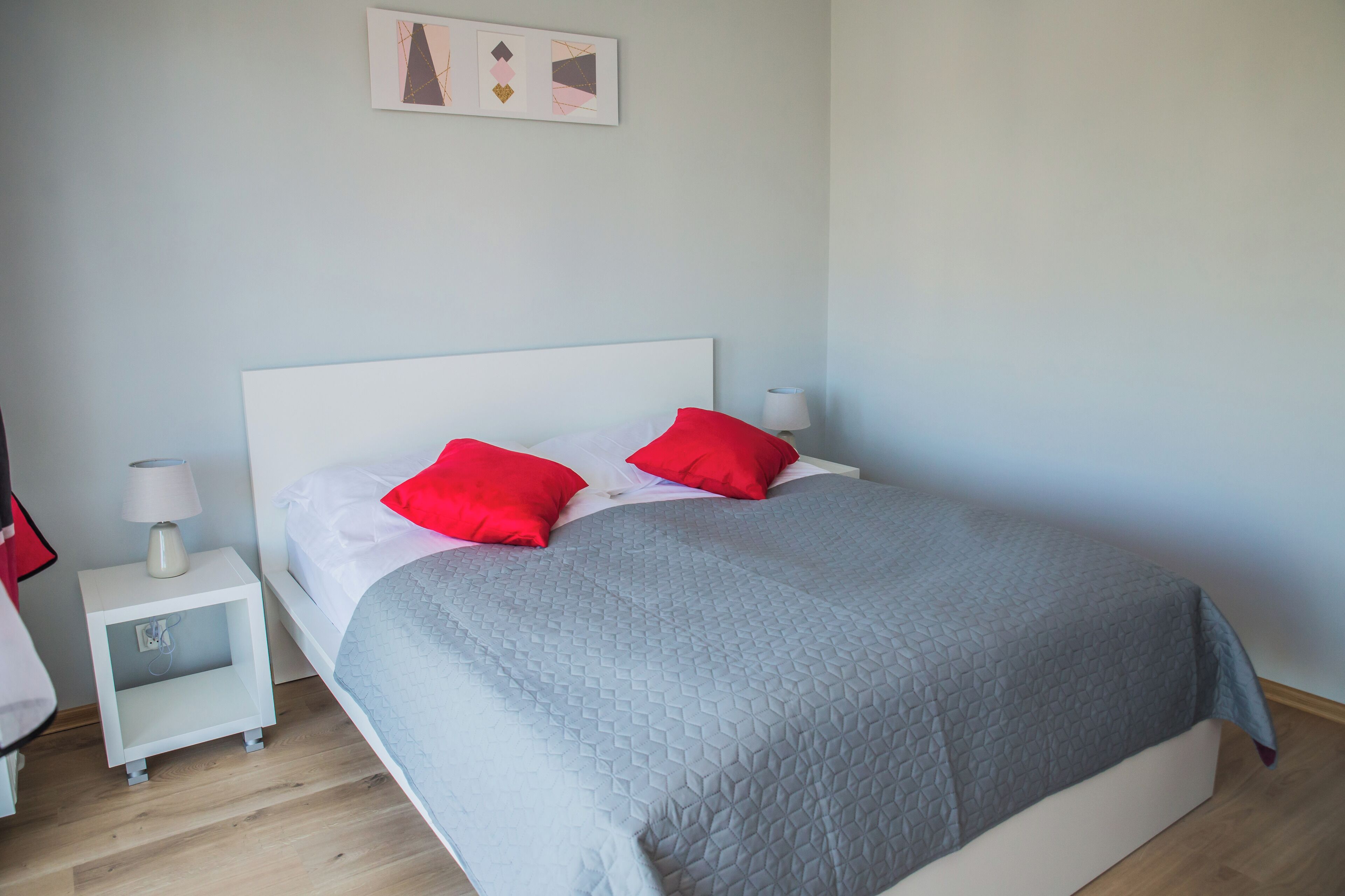 Una cama bien hecha con colcha gris y dos almohadas rojas, una mesita de noche blanca con una lámpara, y un cuadro en la pared.