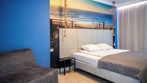 Ein Schlafzimmer mit Bett, einem kleinen Tisch, einem Stuhl und einem Wandbild mit einer Strandansicht.
