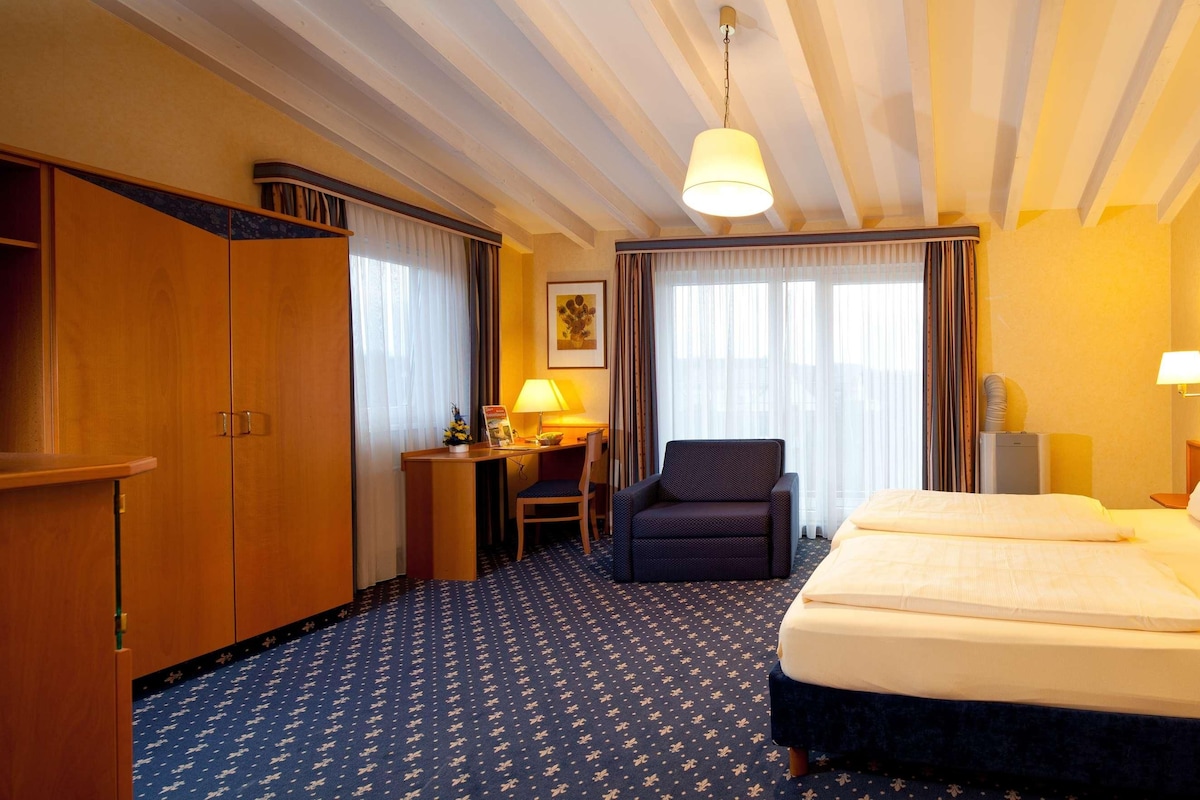 A hotel room with a bed, two bedside lamps, a desk with a chair, a wardrobe, and a large window with curtains.
