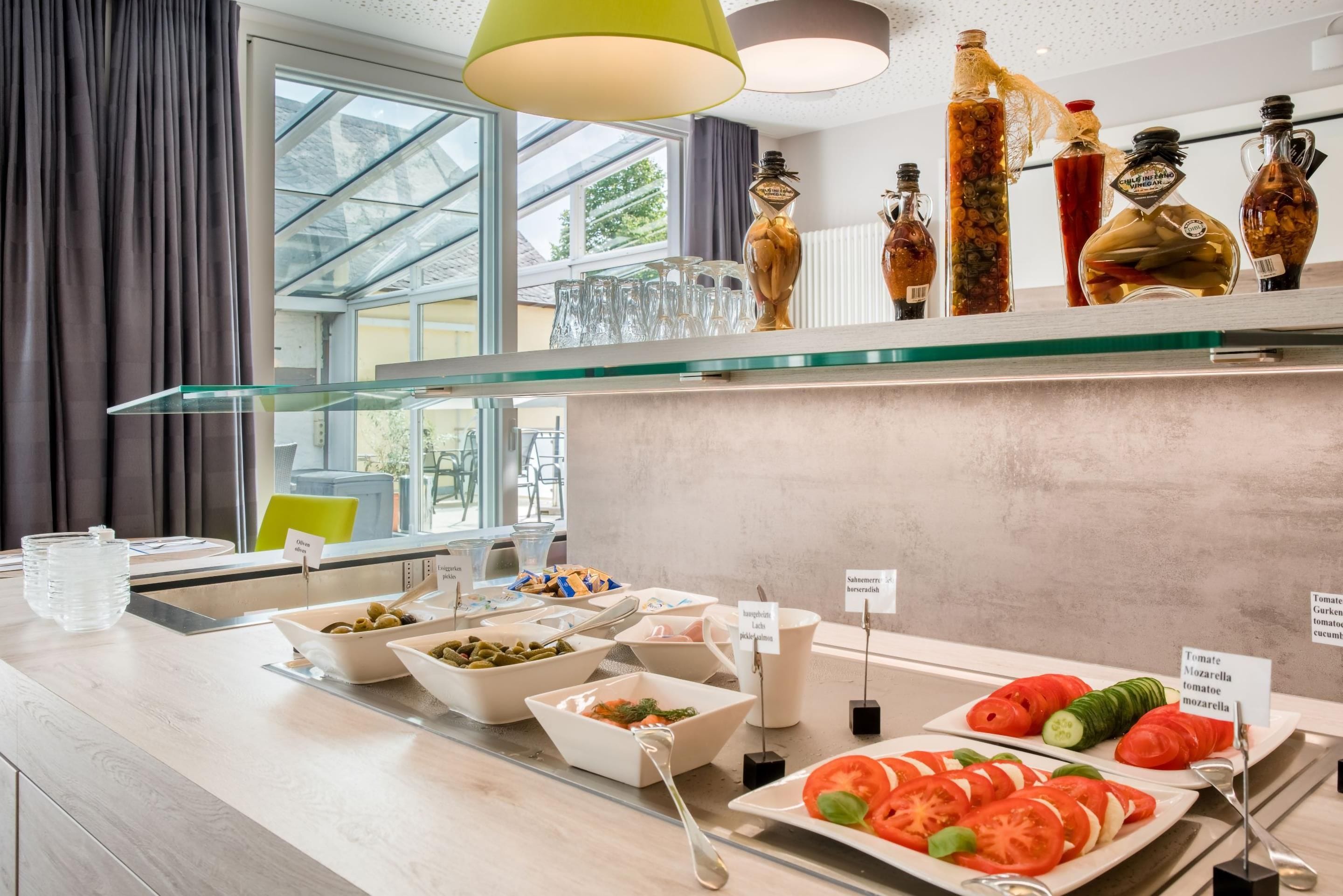 A buffet area with various food options, including olives, pickled cucumbers, and tomato-based dishes.