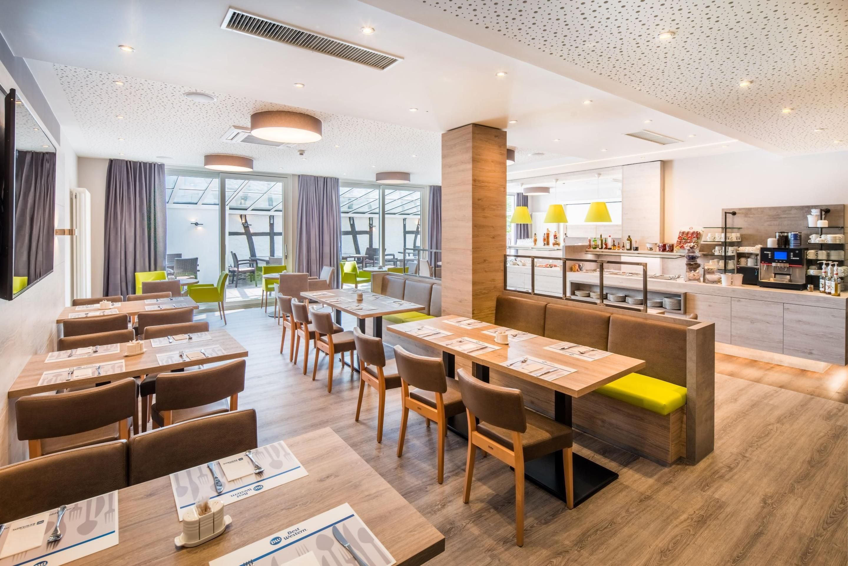 Un comedor moderno con mesas y sillas de madera, una zona de bar con lámparas amarillas y una cocina visible al fondo.
