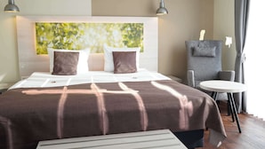A modern hotel room with a large bed, a headboard with a nature-themed artwork, a small round table, and a seating area with a chair.