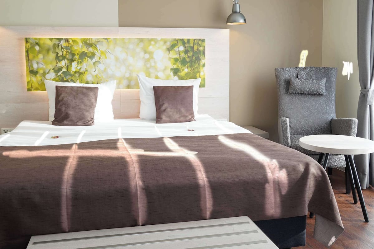 Ein modernes Hotelzimmer mit einem großen Bett, einem Kopfteil mit einem Kunstwerk zum Thema Natur, einem kleinen runden Tisch und einer Sitzecke mit einem Sessel.
