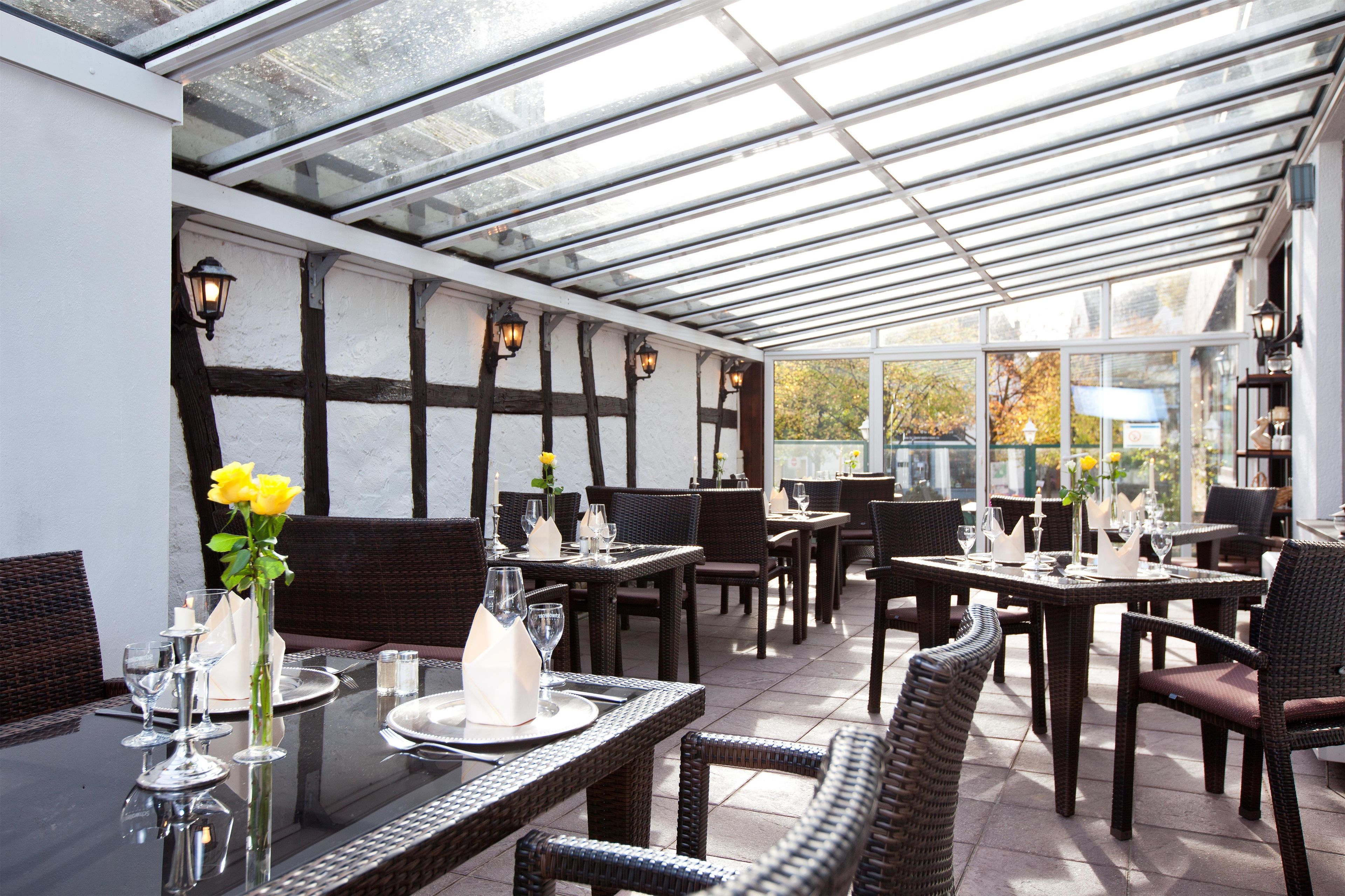 A restaurant with a glass ceiling, wooden beams, and wicker furniture. Tables are set with plates, glasses, and flowers.