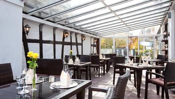 A restaurant with a glass ceiling, wooden beams, and wicker furniture. Tables are set with plates, glasses, and flowers.