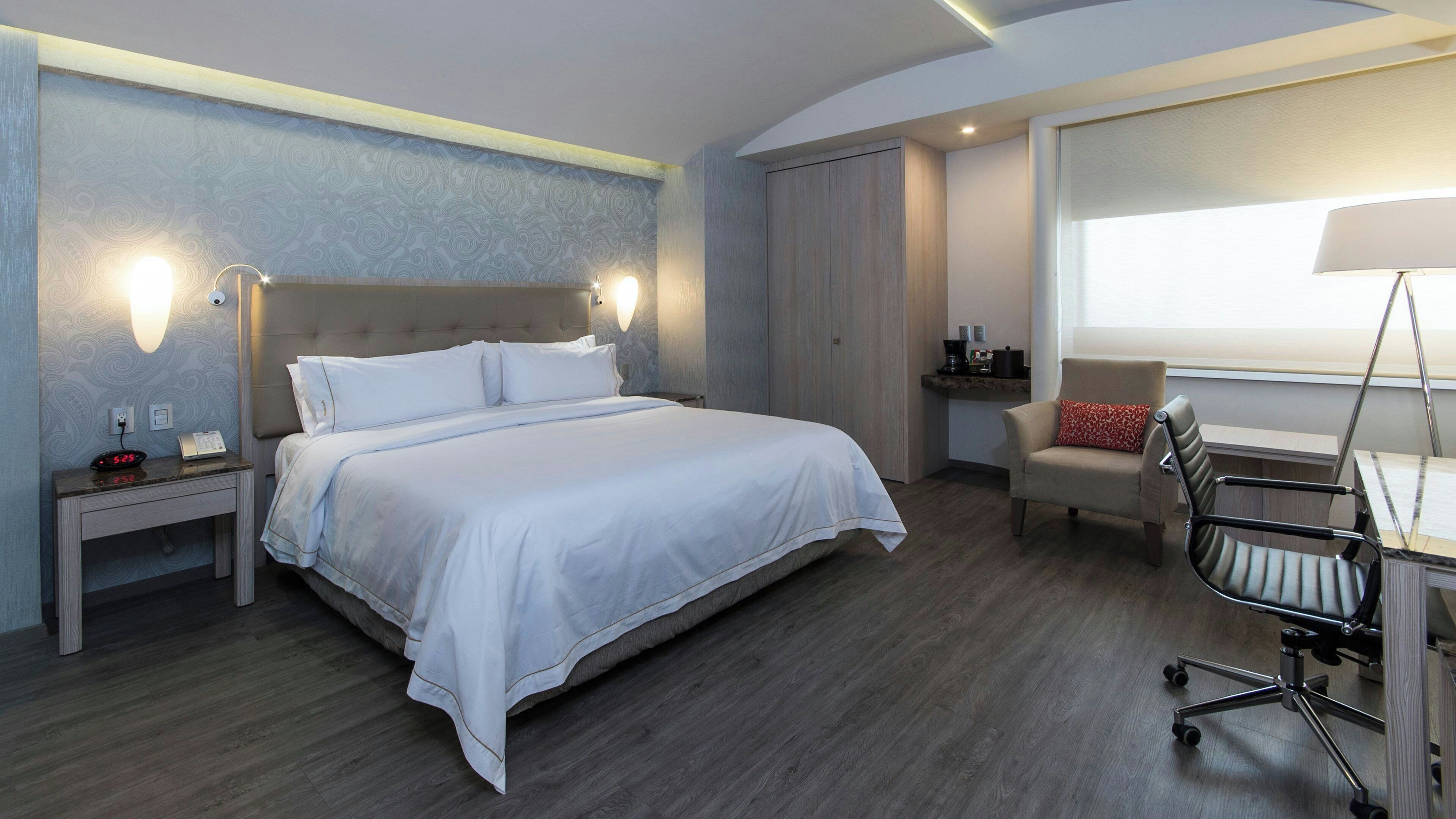 A hotel room with a bed, bedside table, lamp, chair, desk, and a window.