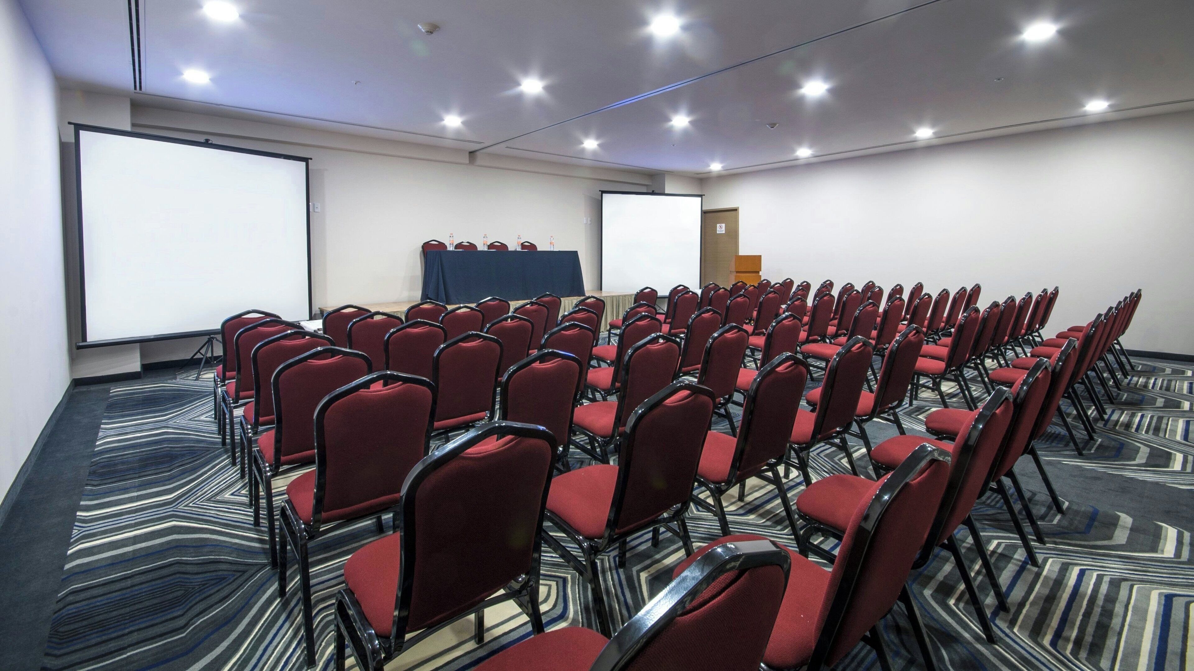 A conference room with rows of red chairs, a projector screen, and a carpeted floor.