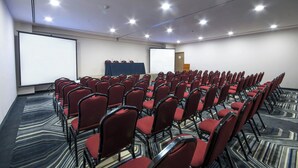 Un salón de conferencias con filas de sillas rojas, una pantalla de proyector y piso alfombrado.