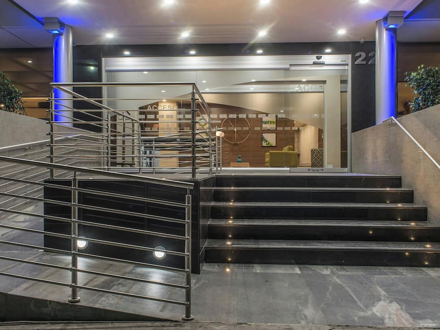 A modern hotel entrance with a glass facade, a metal staircase, and a reception area.