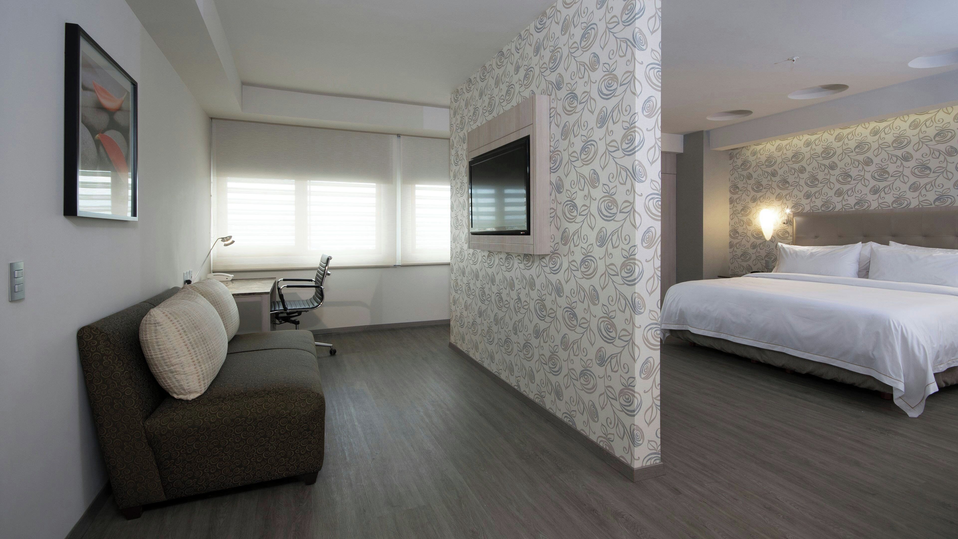 A hotel room with a bed, a sofa, a desk with a chair, a TV mounted on the wall, and patterned wallpaper.