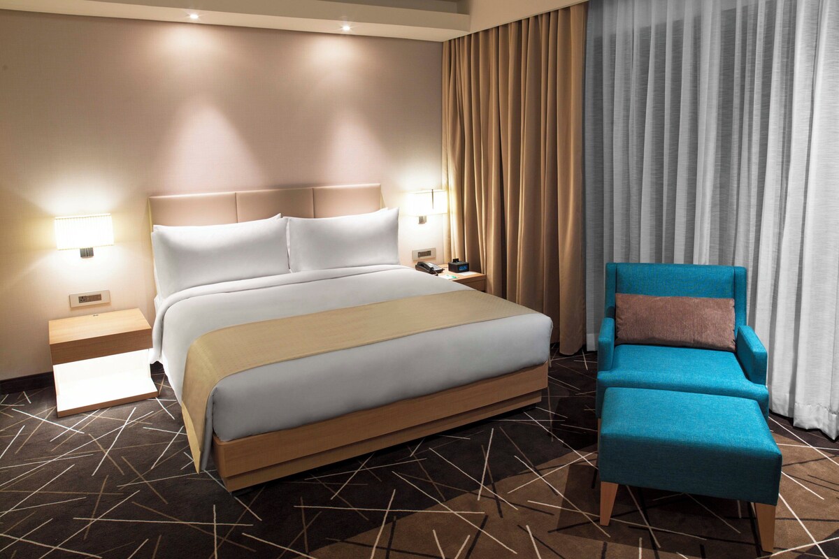 Une chambre d’hôtel avec un grand lit, des lampes de chevet, une table de nuit, un fauteuil bleu et une moquette à motifs.