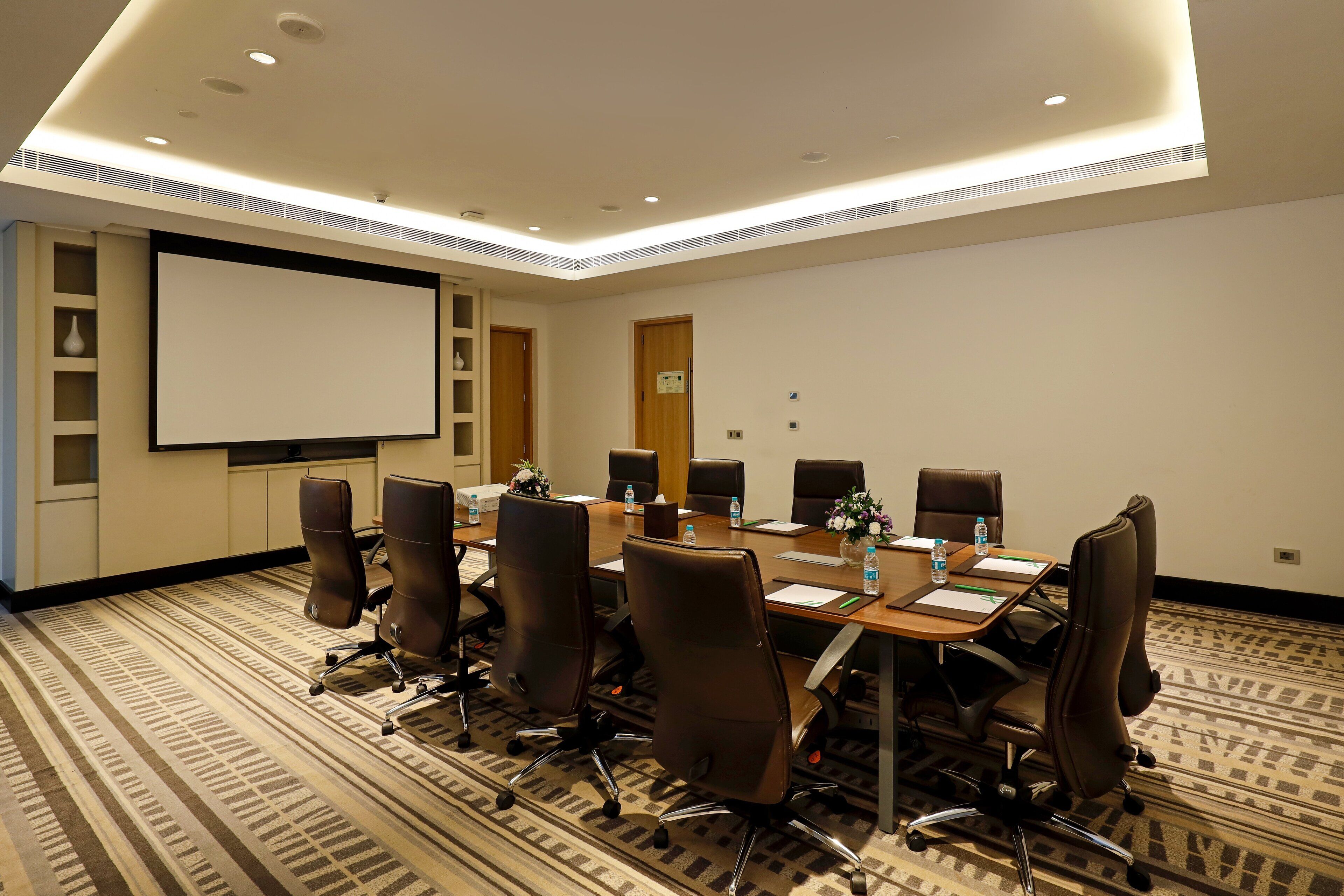 A conference room with a large table, chairs, a projector screen, and a carpeted floor.