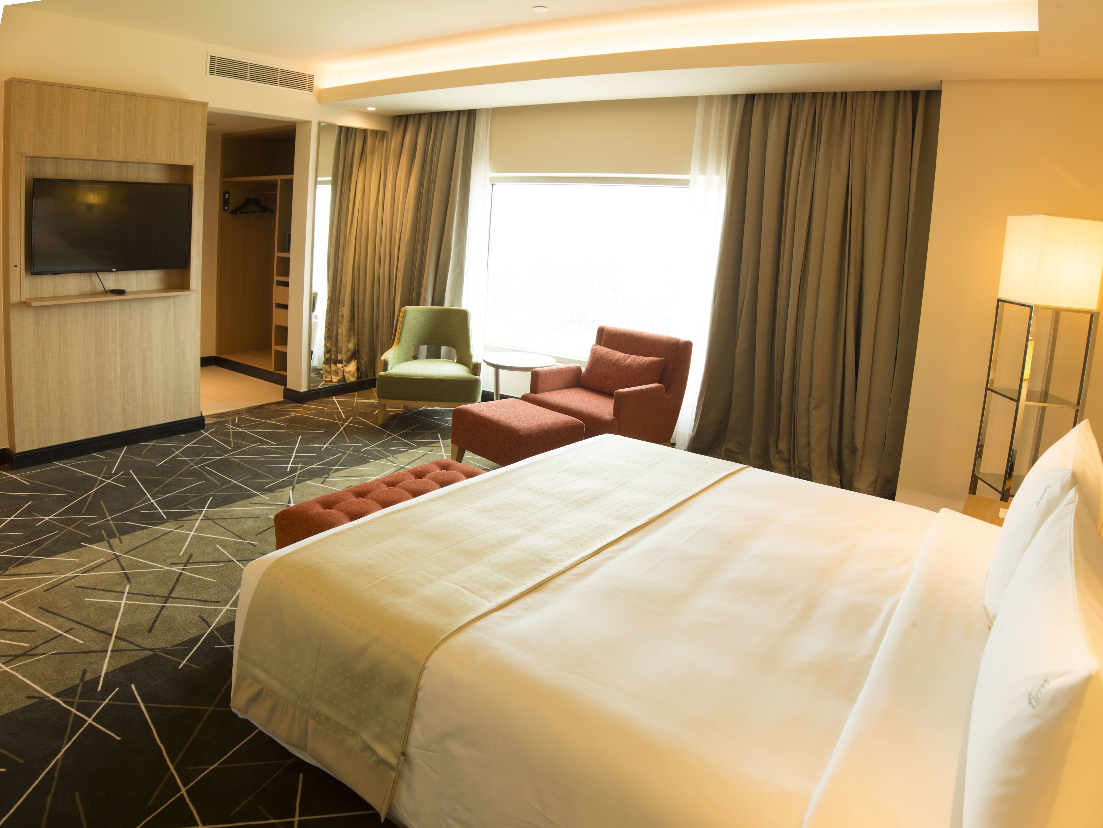 A hotel room with a large bed, a television, a green armchair, a red armchair, and a small table.