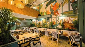 Interior de un restaurante con un mural de loros, mobiliario de mimbre y suelo con estampado.