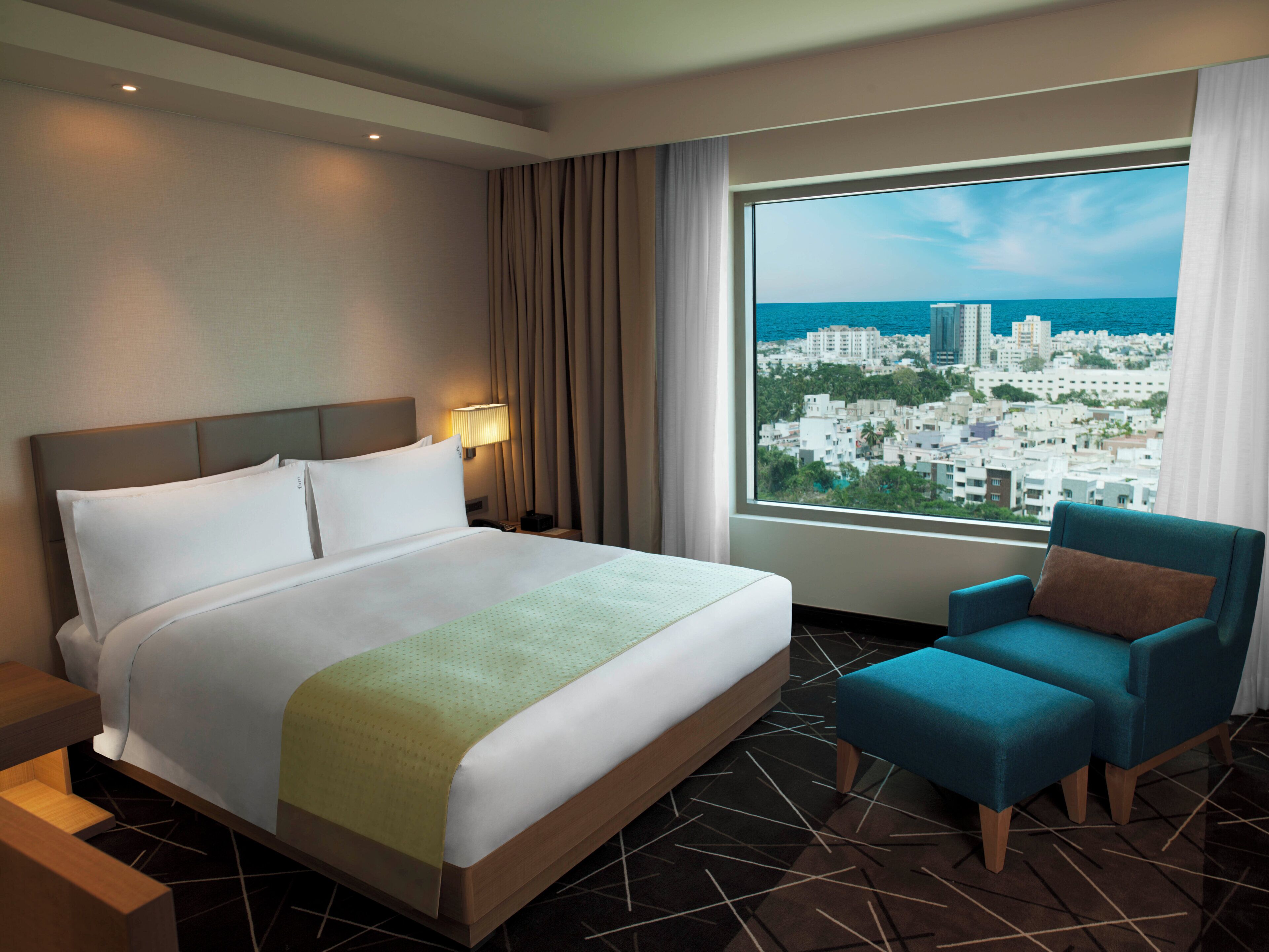 A hotel room with a large bed, a nightstand, a chair, a window with a city view, and a lamp.