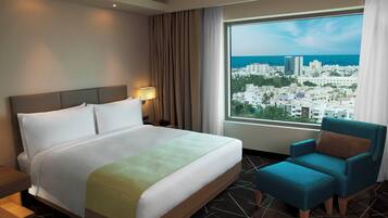 A hotel room with a large bed, a nightstand, a chair, a window with a city view, and a lamp.