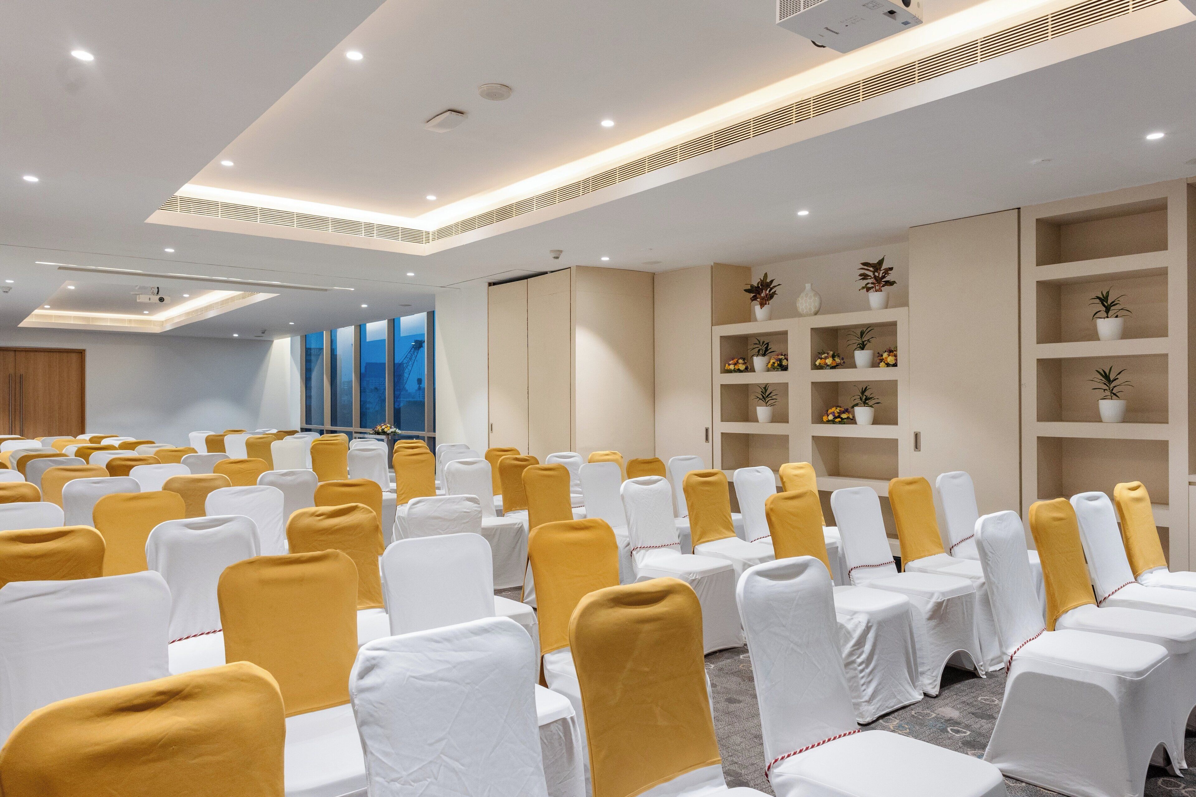 Une salle de conférence avec des rangées de chaises blanches, dont certaines sont ornées de rubans jaunes, et des étagères beiges où sont disposées des plantes en pot.