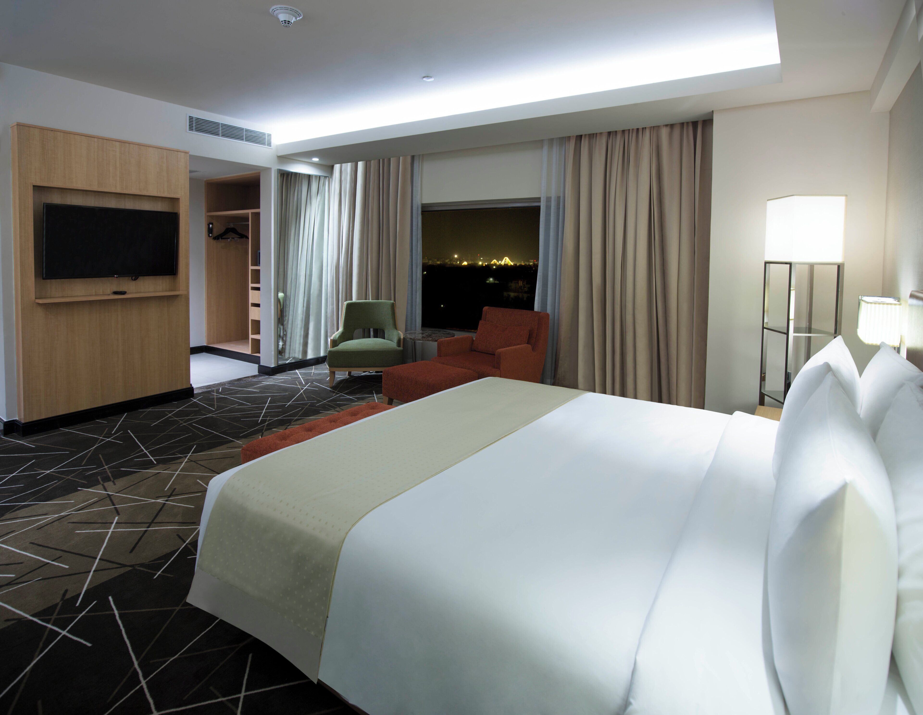 A hotel room with a bed, a television, a seating area with chairs, a view of the cityscape, and a door leading to a balcony.